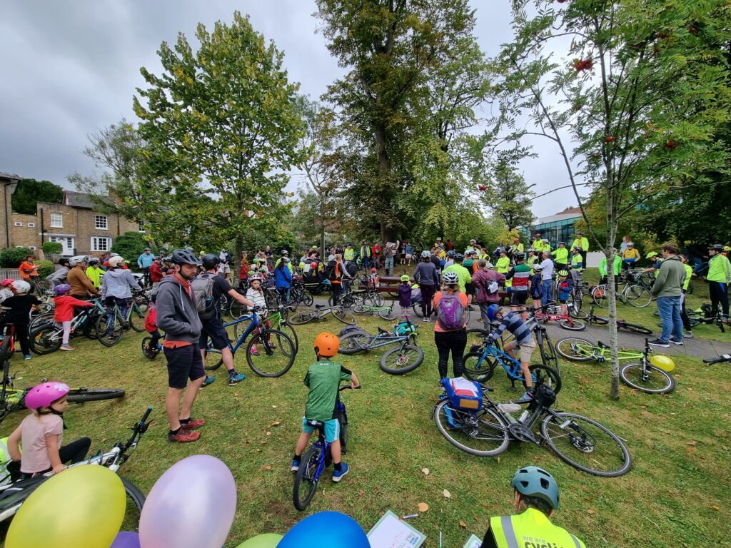 Cyclists congregate before the start of the ride outside GLive. Most people are standing and there are many bikes laying on the grass.