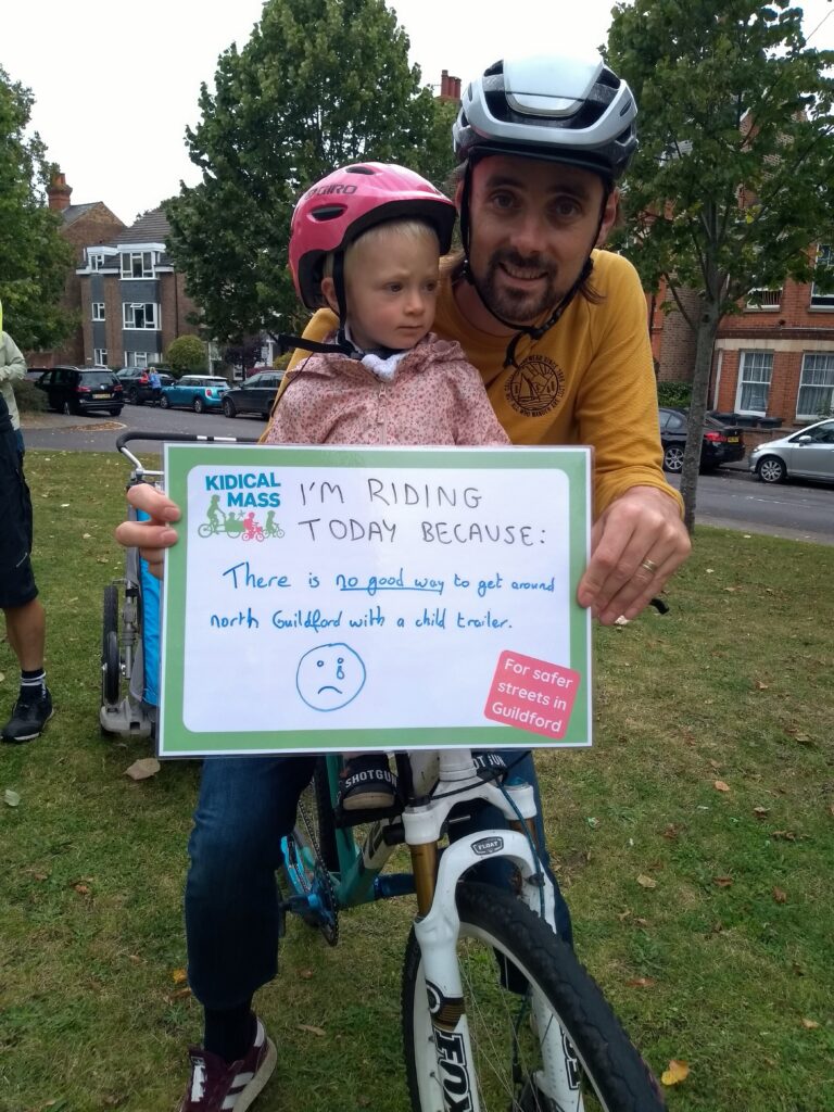 Adult with small child on the front of their bicycle. The adult is holding a sign which reads "There is no good way to get around north Guildford with a child trailer."