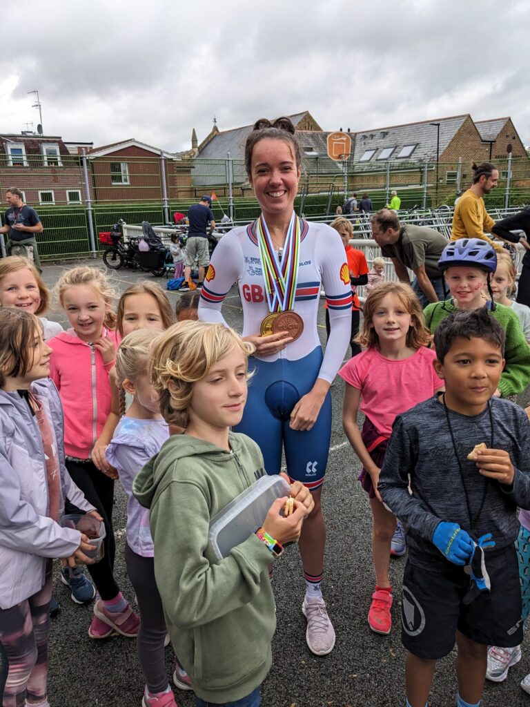 Lizzi Jordan wearing her Gold and Bronze medals surrounded by children looking happy, one child is eating a biscuit